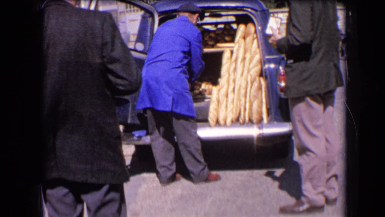 Baker in a blue coat reaches into the open back of a blue van filled with baguettes.