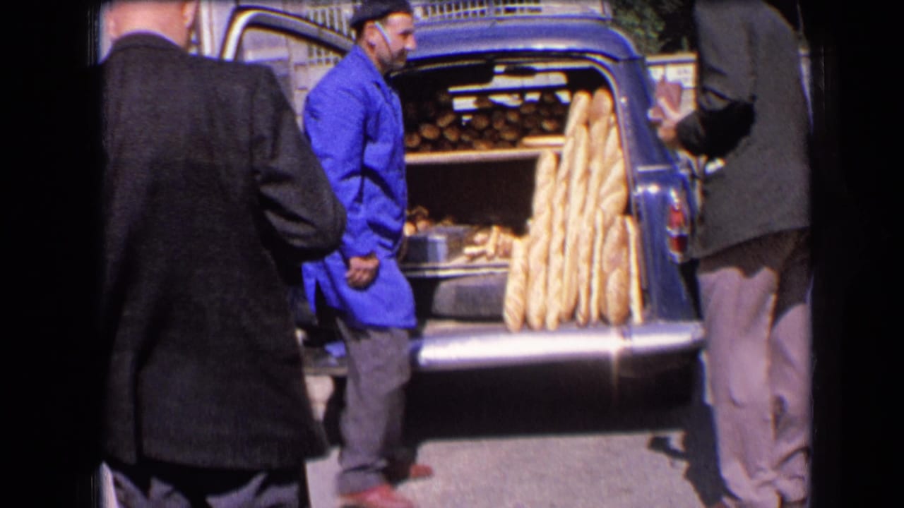Man in a blue coat stands beside an open blue van with baguettes arranged inside.