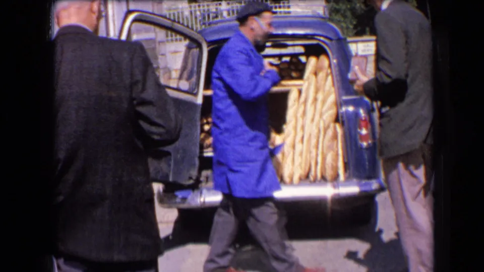The baker moves across the frame near a blue van loaded with long baguettes and smaller loaves, with suited men in the foreground.