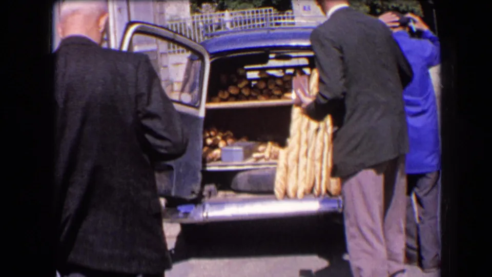 A man in a dark jacket handles bread at the open rear of a blue van in France, 1963, while the baker in a blue coat stands at the side.