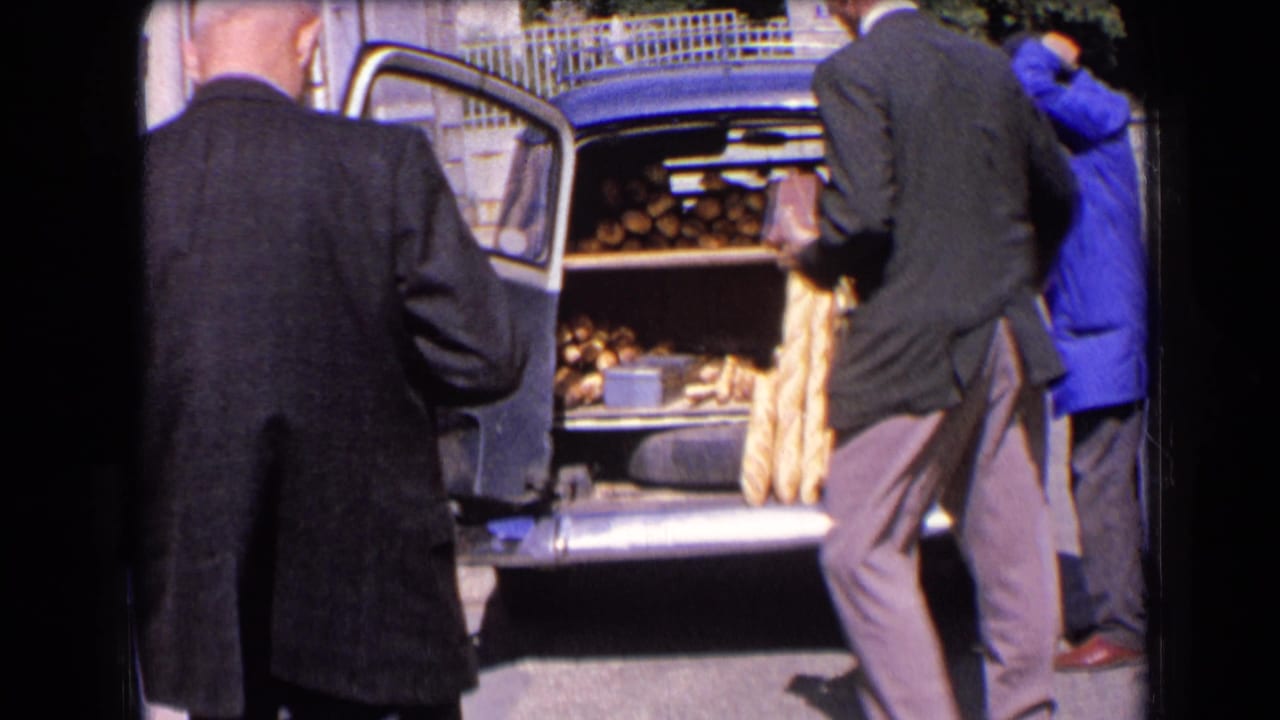 1963 France home-movie frame showing a baker in a blue coat loading baguettes into a vintage blue car trunk while suited adults watch.