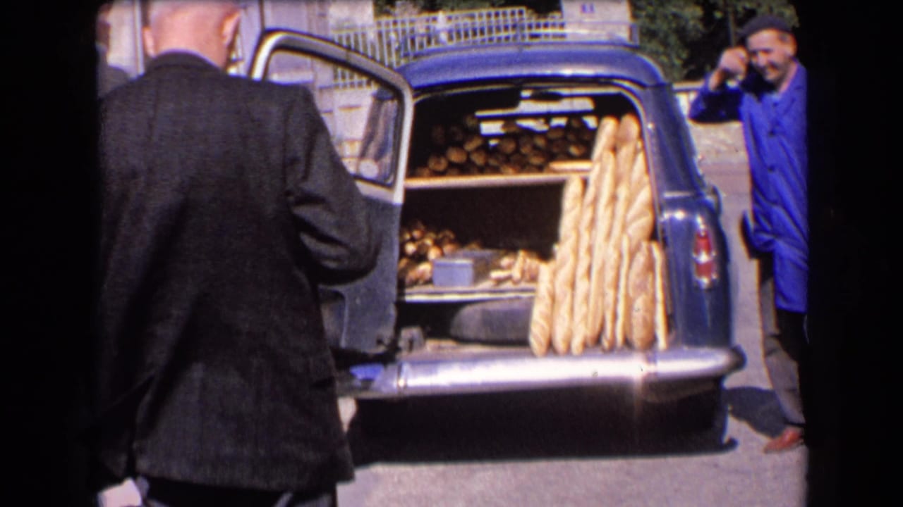 Open trunk of a blue car filled with bread, with a baker standing at the right edge.