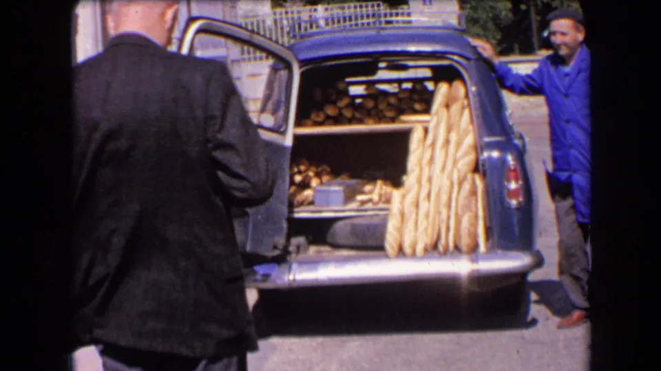 1963 France home-movie frame showing a suited customer near a blue car trunk loaded with baguettes and round loaves.
