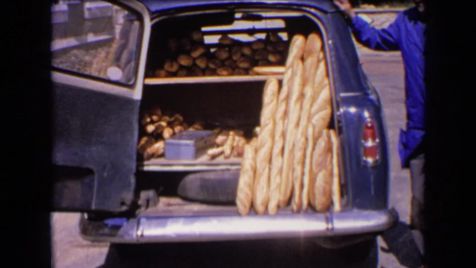 1963 France home-movie frame of a vintage car opened to show shelves of bread, with baguettes leaning near the rear bumper.