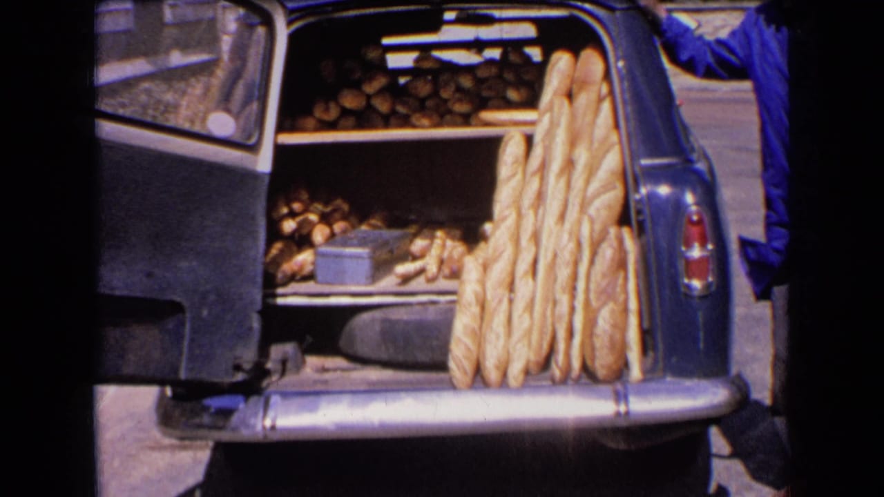 The open rear of the car shows stacked round loaves and upright baguettes, with part of a blue-smocked person at the side.