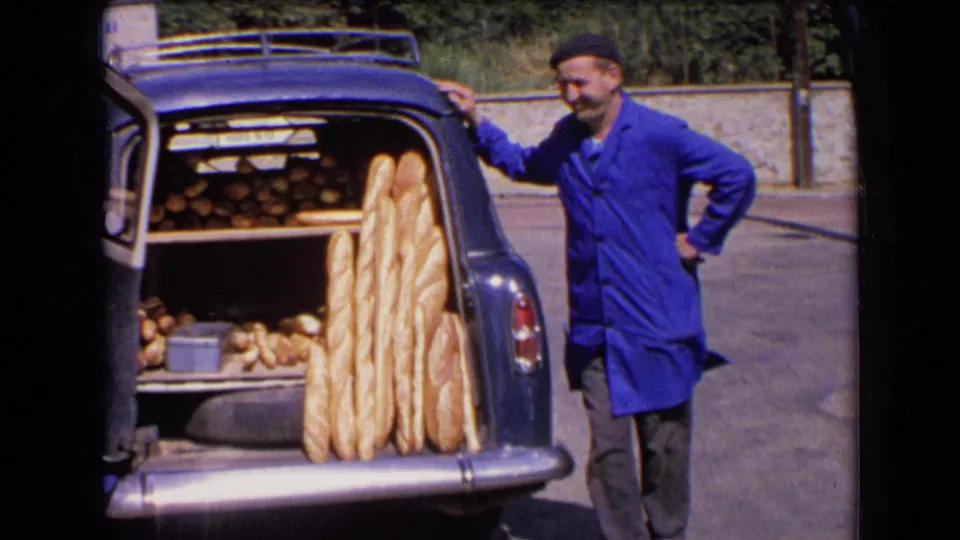 A baker in a blue smock and dark cap stands beside the open rear of a vintage car loaded with baguettes and loaves in France, 1963.
