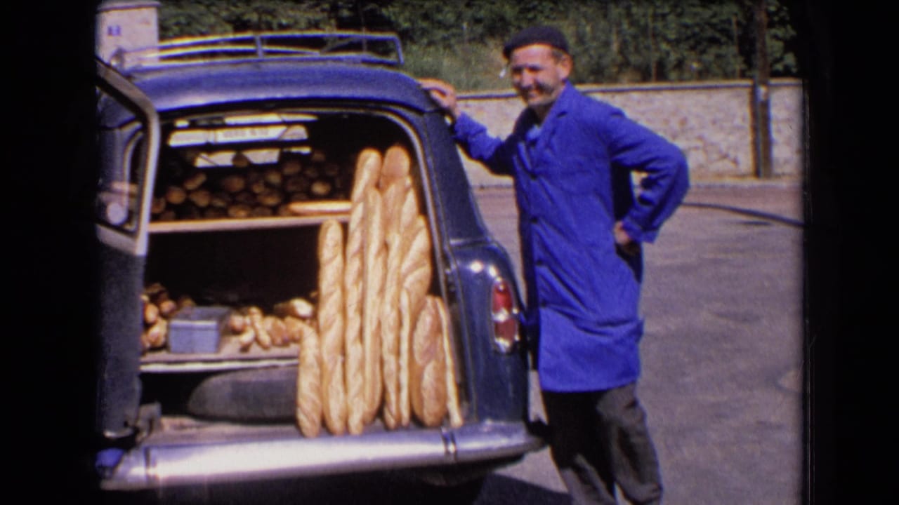 In France, 1963, a baker in a blue coat and beret smiles beside a vintage car trunk packed with baguettes and smaller loaves.