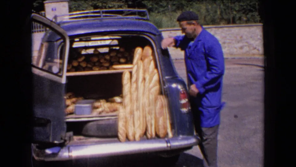 The baker turns toward the bread display in the vintage car trunk, with long baguettes and smaller loaves visible on shelves.