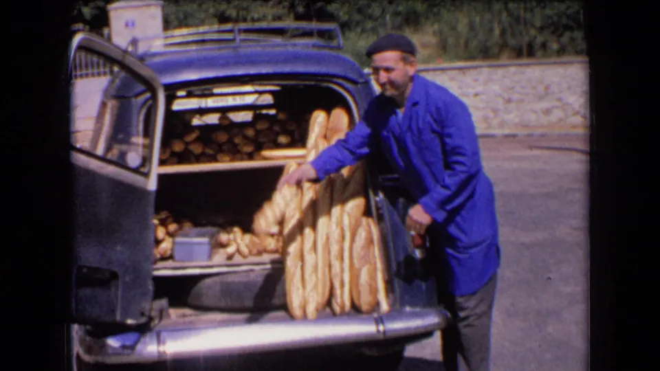 The baker leans into the open trunk and reaches toward the baguettes, with smaller loaves visible behind them in the car.