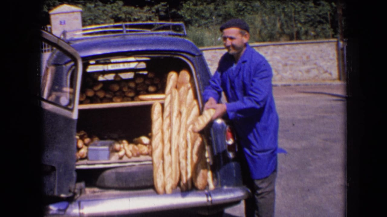 Baker in a blue coat loads long baguettes into the open trunk of a blue car.