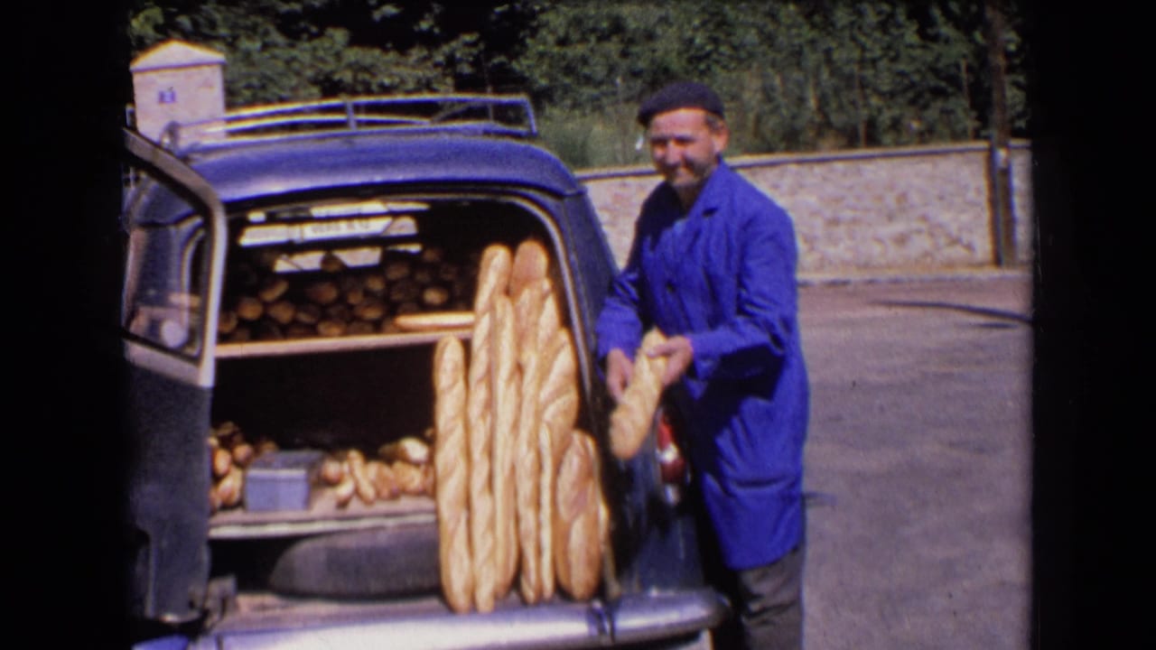 The smiling baker holds a loaf near a car trunk packed with baguettes and other bread in 1963 France.
