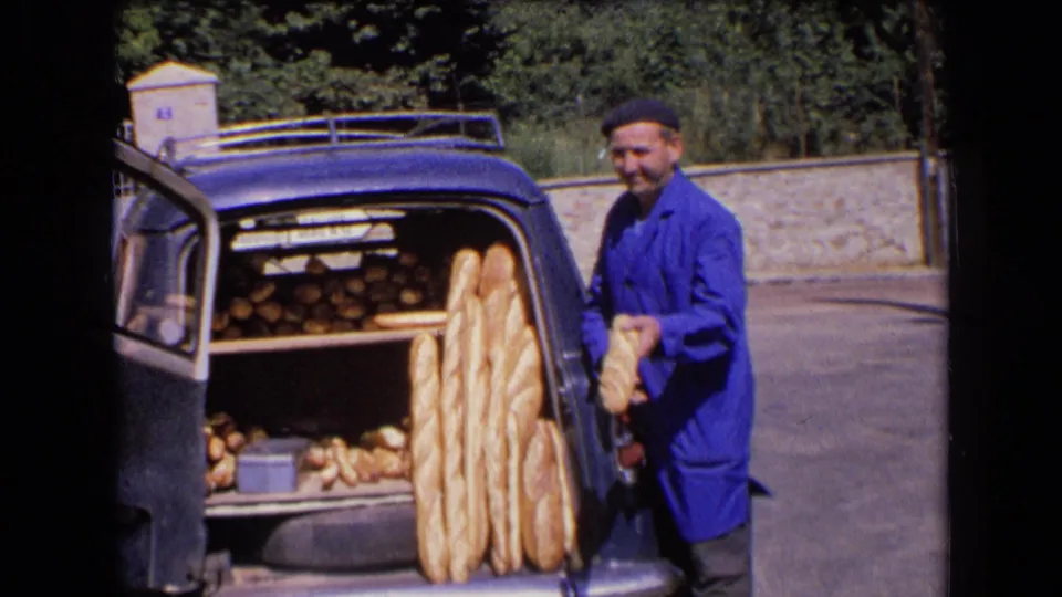 A baker in work clothes holds bread beside the open trunk, where baguettes and smaller loaves are arranged for delivery.