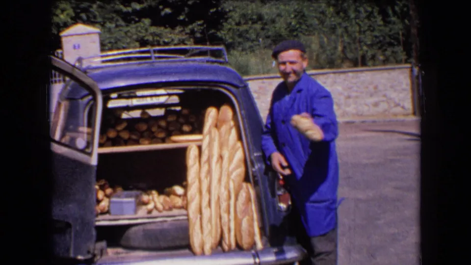 The baker faces the camera with bread in hand beside a blue car used as a mobile bakery in 1963 France.