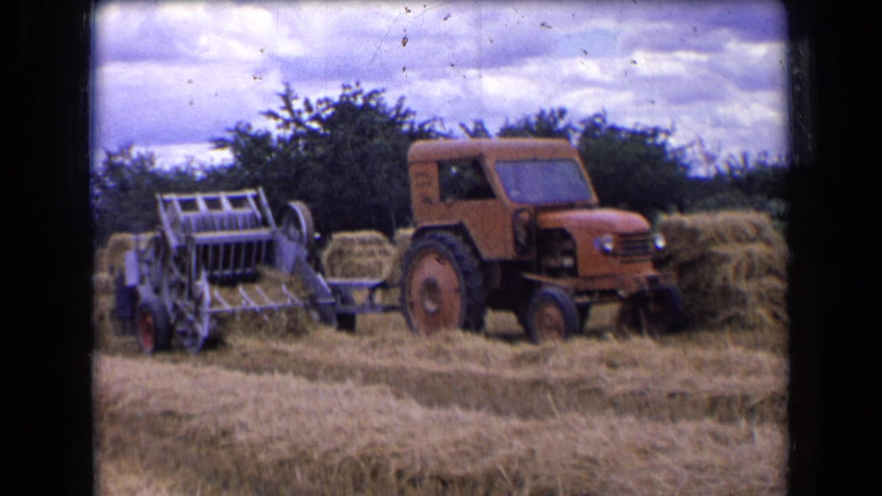 1963 rural France 8mm frame showing an orange tractor with a driver pulling a hay baler through cut hay.
