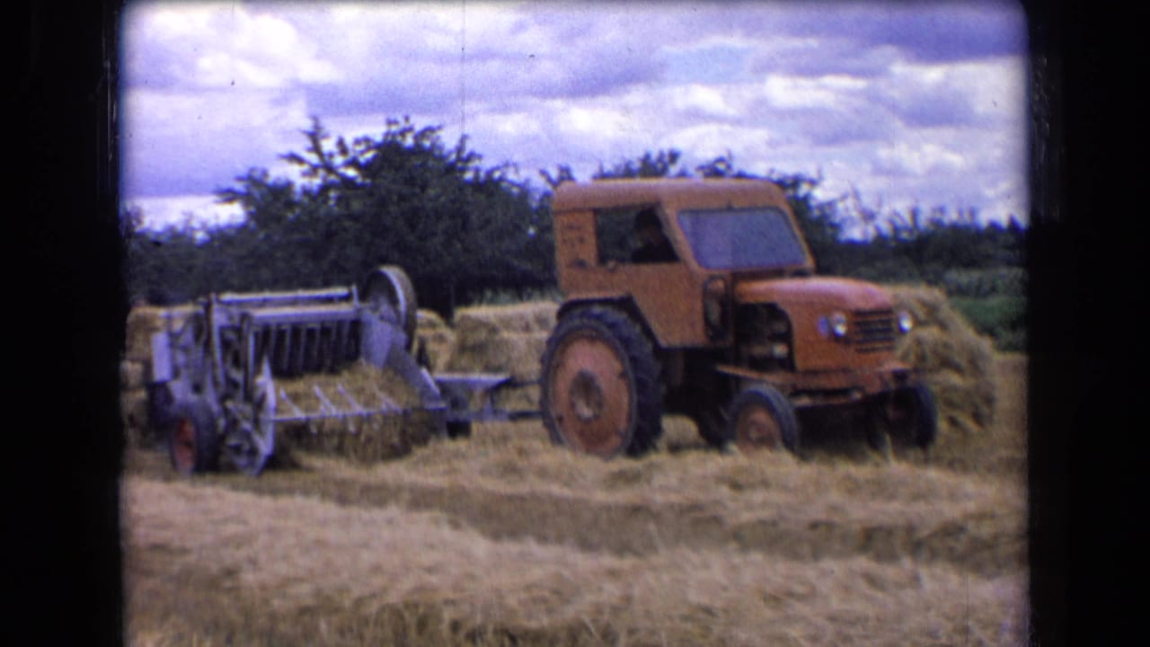 1963 rural France 8mm frame of a tractor towing a baler past hay rows and bales near a tree line.