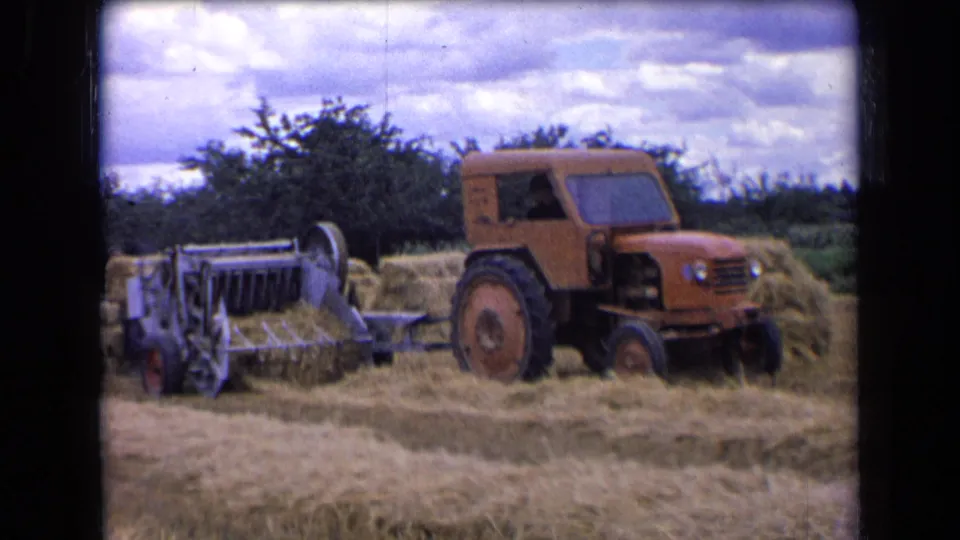 1963 rural France 8mm frame of a tractor towing a baler past hay rows and bales near a tree line.