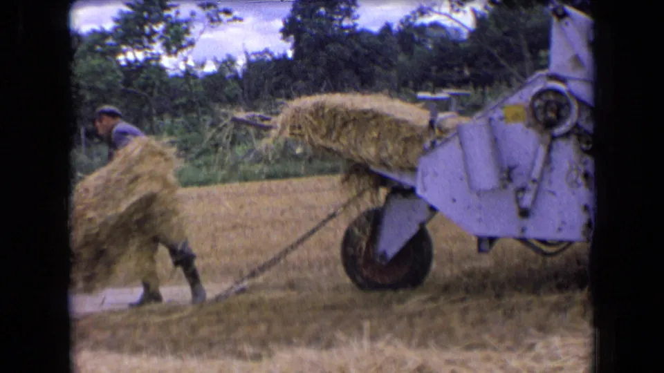 Close 8mm view of hay entering a gray baler while a worker moves bales in a rural French field.