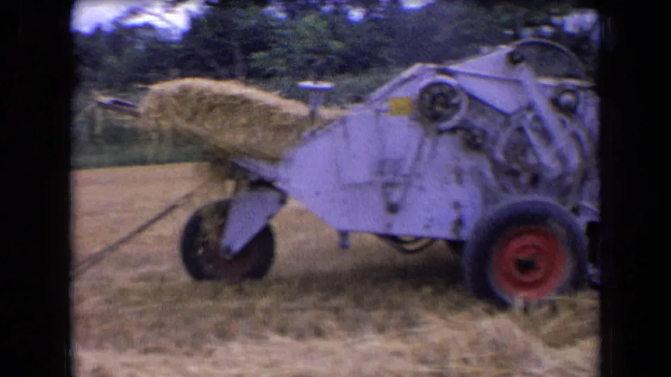 The gray hay baler pushes out a formed bale during 1963 farm work in France, with trees beyond the field.