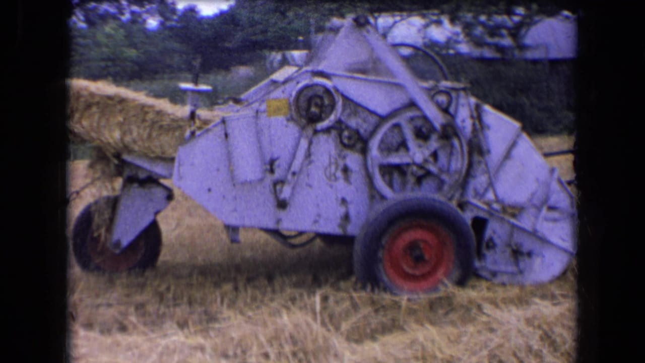 1963 France 8mm frame of a light-colored hay baler with red wheels pushing a bale from its rear chute in a harvested field.