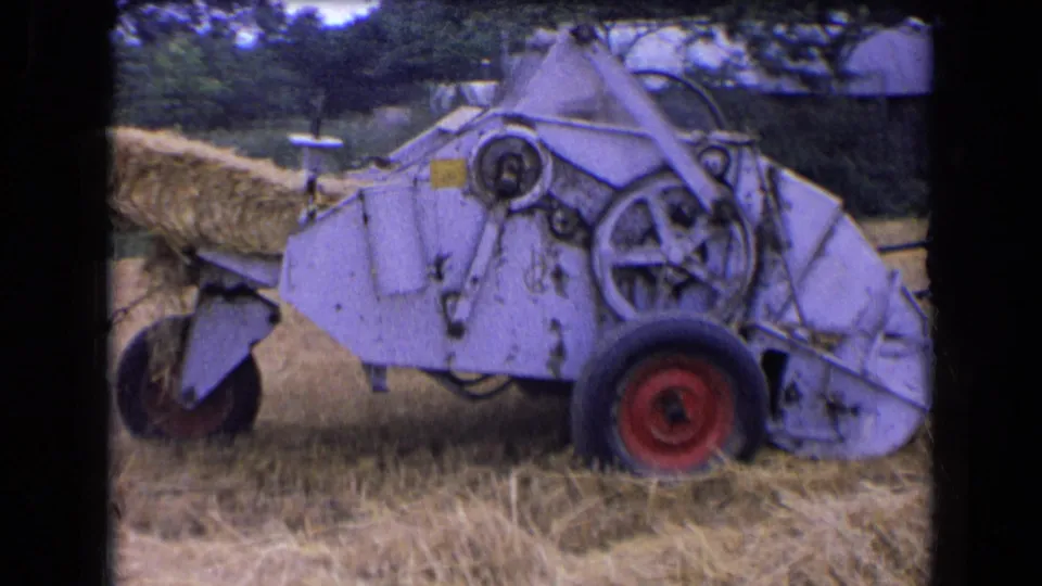 1963 France 8mm frame of a light-colored hay baler with red wheels pushing a bale from its rear chute in a harvested field.