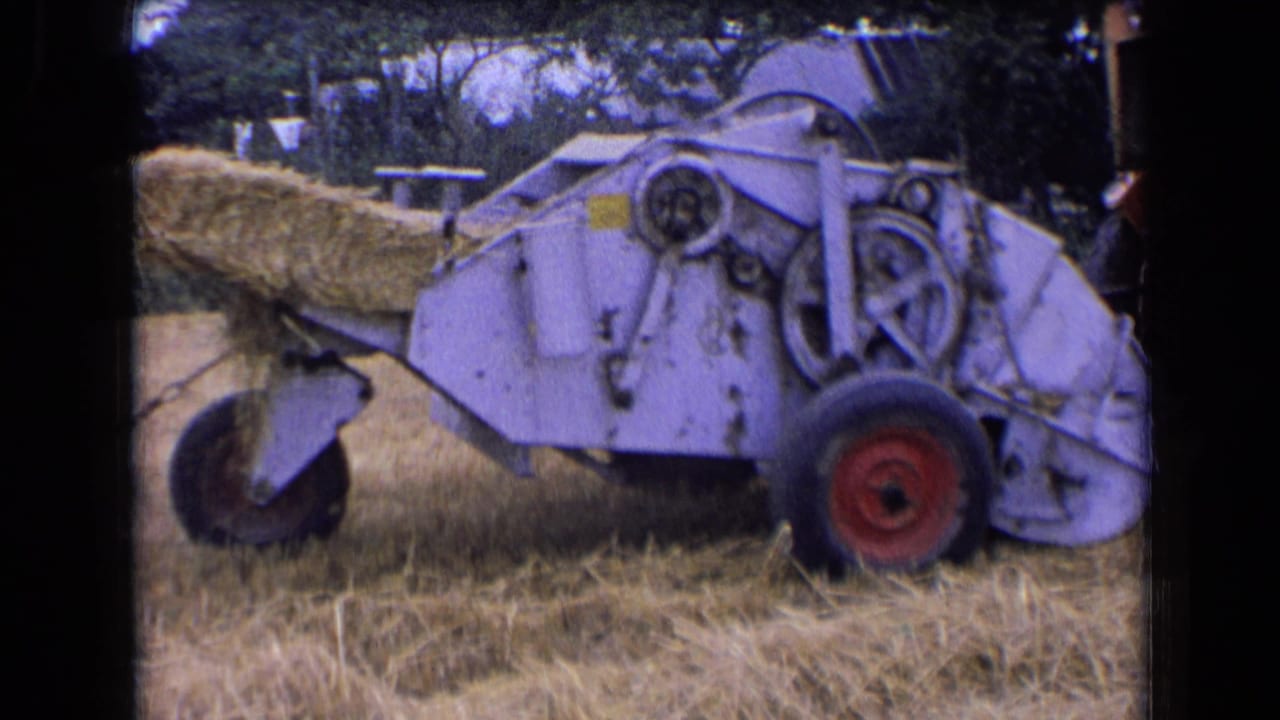 1963 France 8mm frame showing the side of a hay baler, its exposed mechanical parts, red wheel, and hay bale exiting behind.