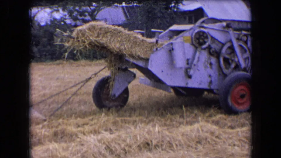 1963 France 8mm frame of the baler from a closer side angle, showing the red wheel, metal housing, and hay moving through the chute.