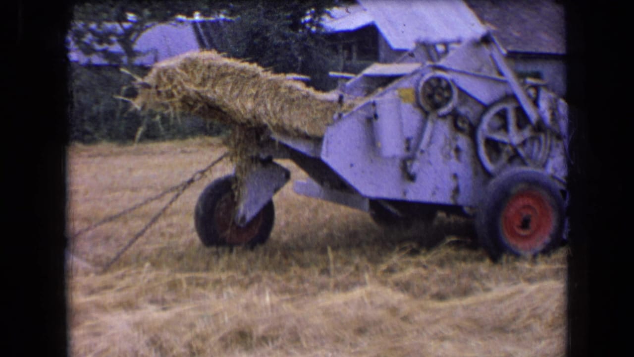 1963 France 8mm frame showing a white baler carrying straw in a cut field, with red wheels and rural buildings behind it.