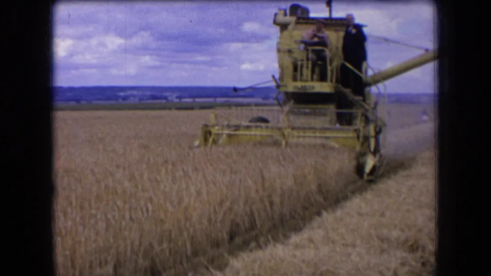 1963 France 8mm frame showing a combine moving across a wheat field with two adult workers visible against a cloudy horizon.