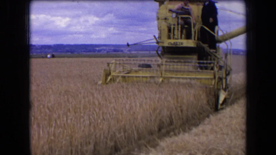 1963 France 8mm frame of a combine's cutting header moving into standing wheat, with two men riding above it.