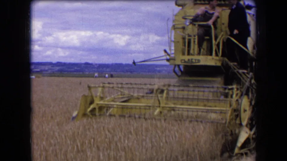 The yellow combine continues across the wheat field in France, with an operator seated and another man standing nearby.
