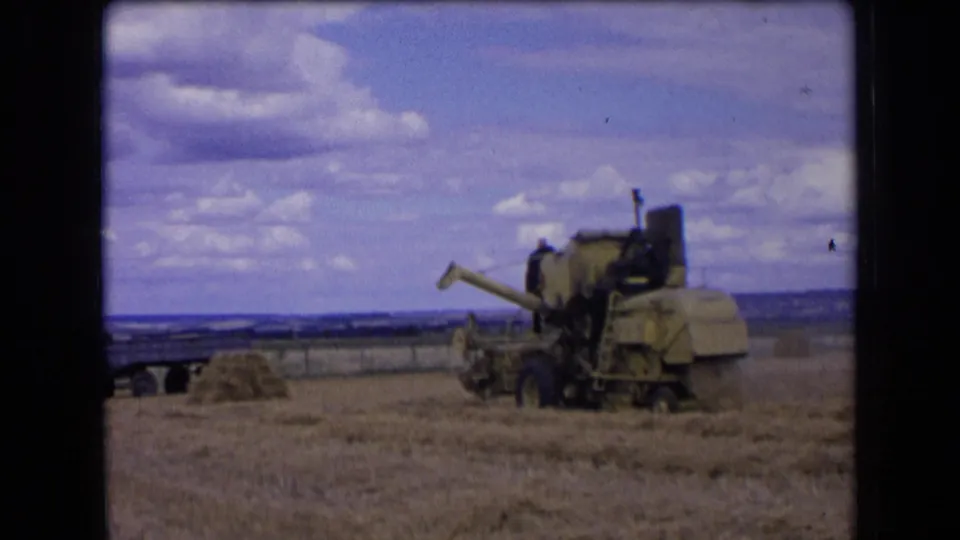 A combine harvester appears farther across the French wheat field, framed by stubble, straw piles, and a broad clouded sky.