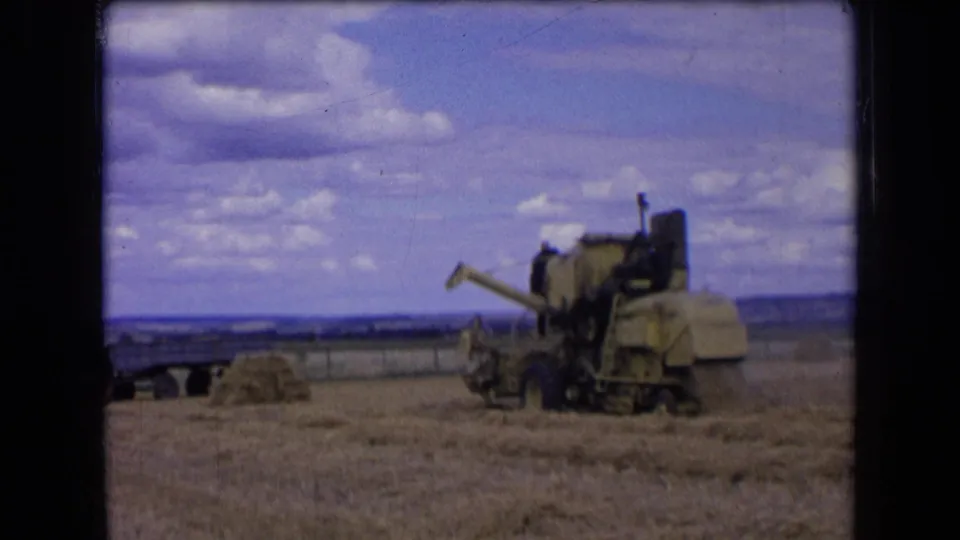 1963 France 8mm frame showing a combine harvester in a harvested wheat field with straw piles, a trailer, and cloudy sky.
