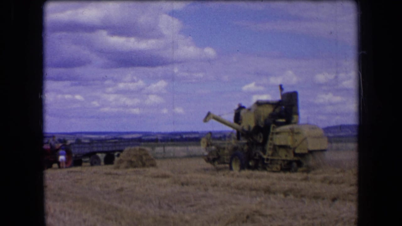 1963 France 8mm frame of a combine harvester in a wheat field, with small people near the machine and a wagon in the distance.