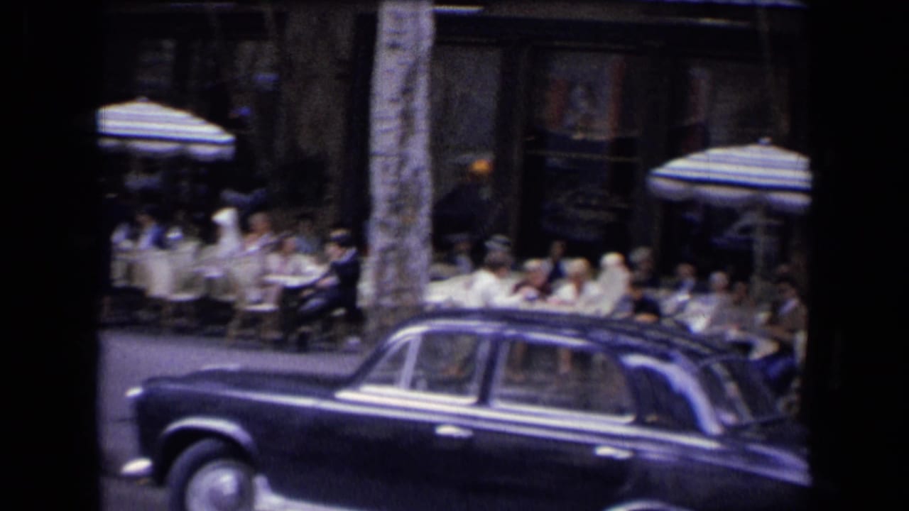A dark mid-century sedan passes a busy French outdoor restaurant with seated diners and striped umbrellas in a 1963 home-movie frame.