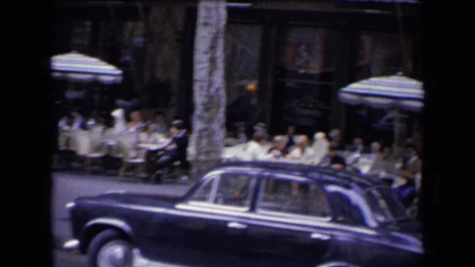 A dark mid-century sedan passes a busy French outdoor restaurant with seated diners and striped umbrellas in a 1963 home-movie frame.