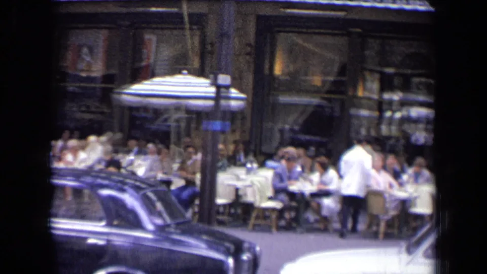 French street restaurant scene with diners, waiters in light jackets, striped umbrellas, and parked or passing cars, filmed on 8mm in 1963.