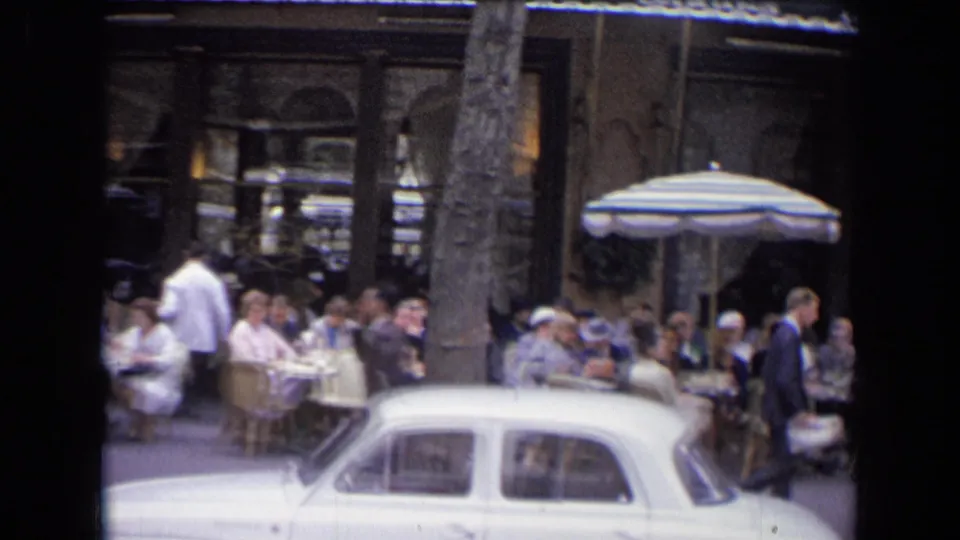 A white compact car crosses the foreground of a French outdoor restaurant scene with pedestrians, diners, umbrellas, and storefronts visible.