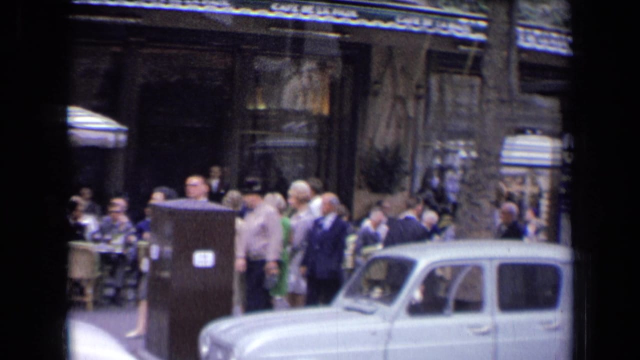 Street-level 1963 France view of a crowded cafe frontage, with pedestrians in period clothing and a white vintage car at the curb.