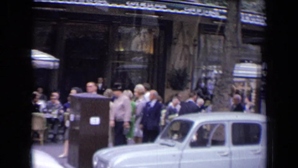 Street-level 1963 France view of a crowded cafe frontage, with pedestrians in period clothing and a white vintage car at the curb.