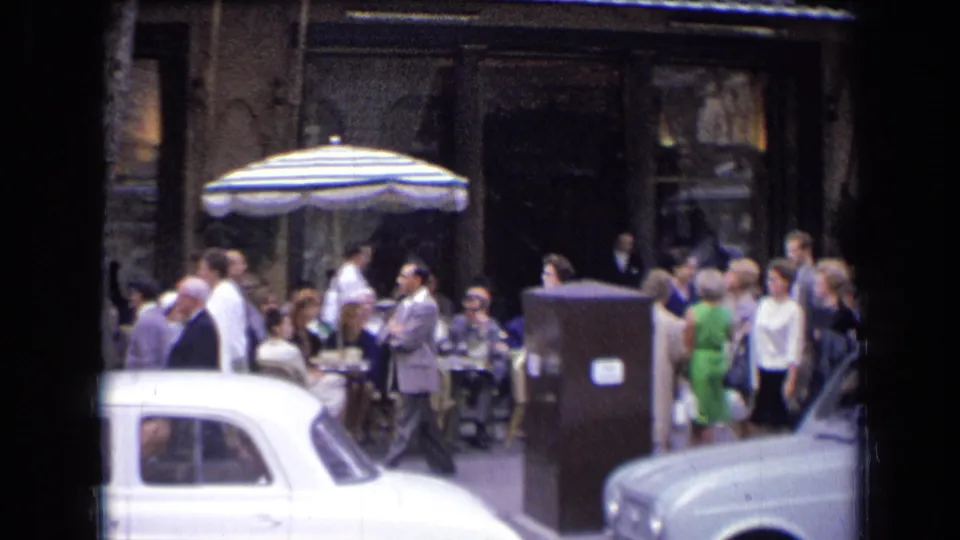 1963 France cafe view with diners under a striped umbrella, pedestrians in suits and dresses, and white vintage cars along the street.