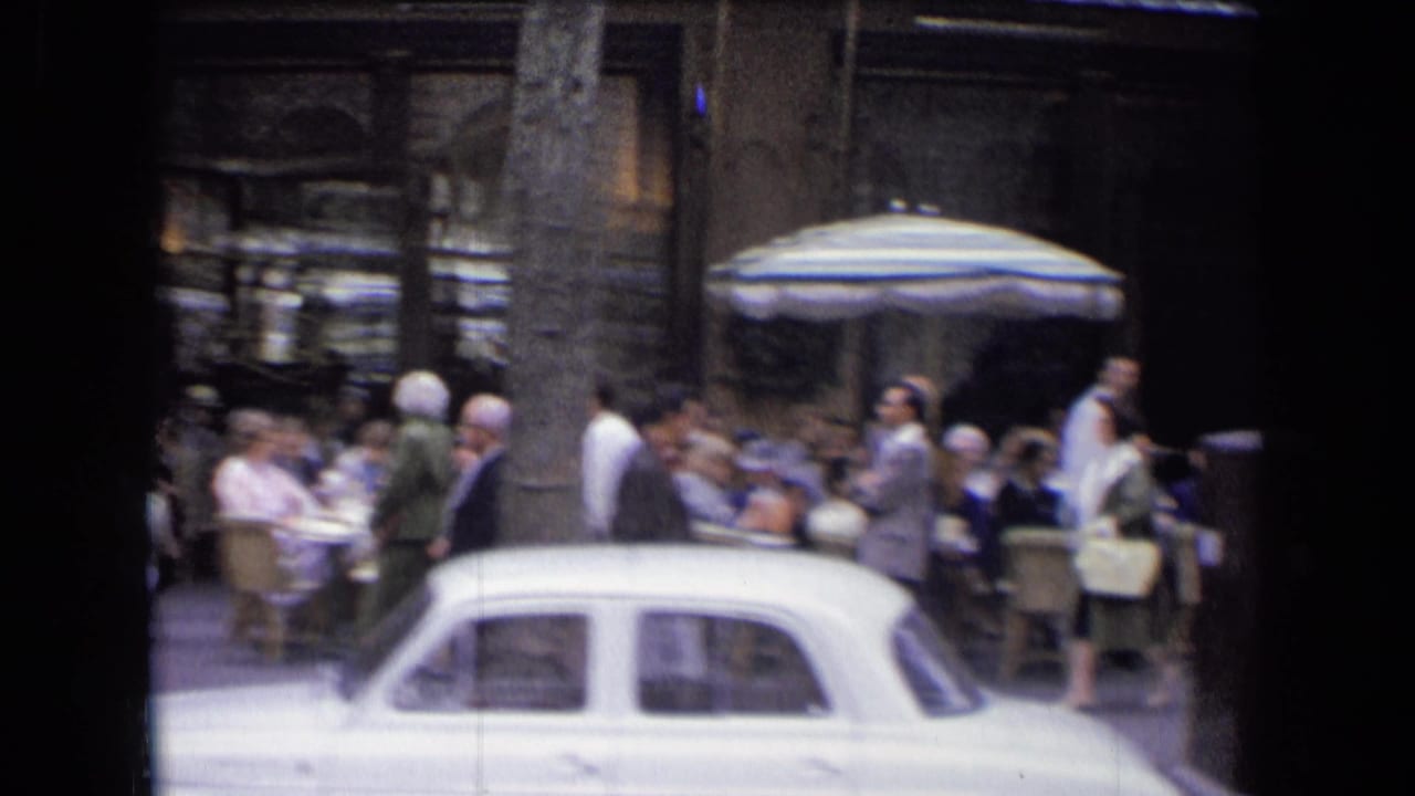 1963 France street view with café patrons under a striped umbrella as a white sedan passes in the foreground.