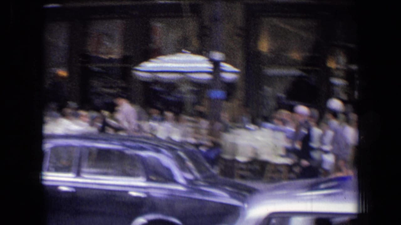 Dark sedan passing café seating with pedestrians and umbrellas behind it.
