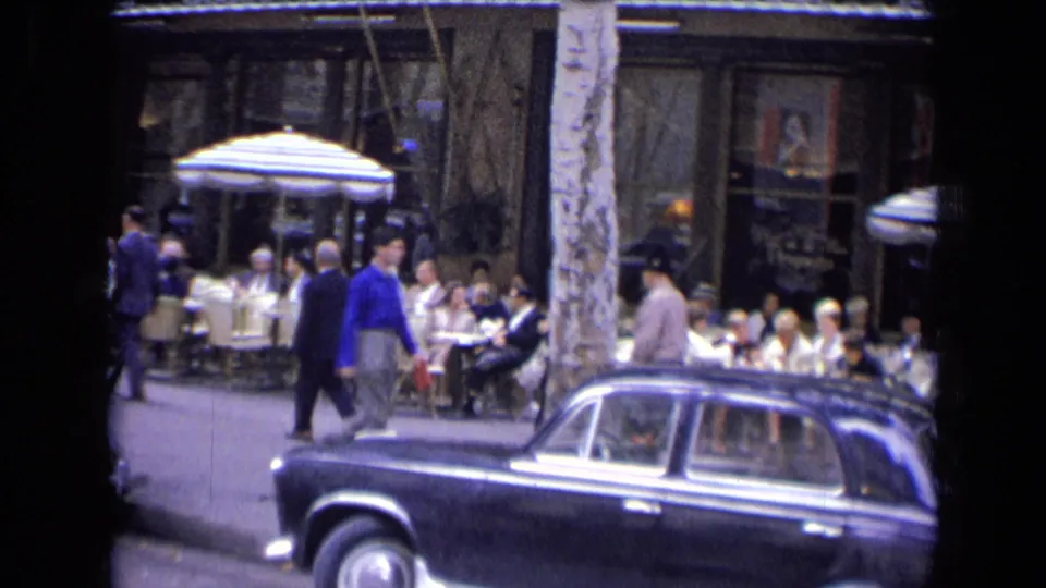 Street-level 8mm view in 1963 France with pedestrians near café seating and a dark mid-century car at the curb.