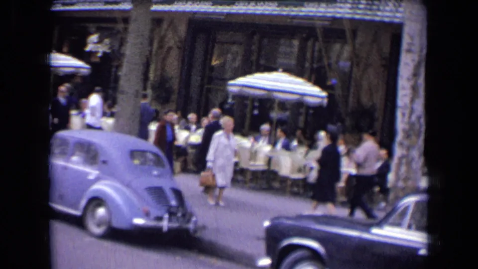 1963 France street view with pedestrians, outdoor café seating, a pale vintage car, and a dark sedan at the frame edge.