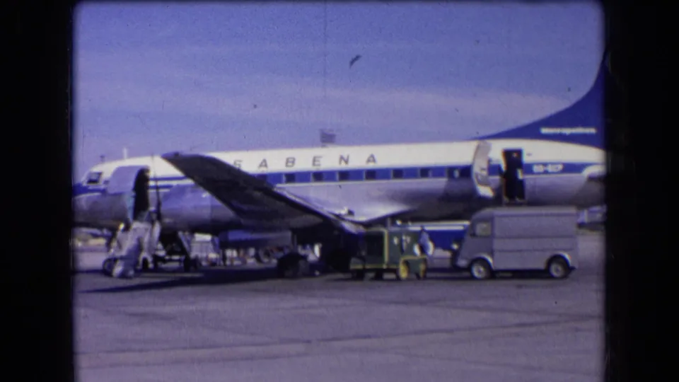 1963 France 8mm frame showing a Sabena aircraft parked on the tarmac with its door open, stairs deployed, and ground vehicles nearby.