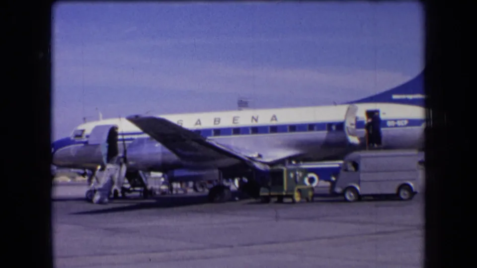 1963 France frame of a blue and white Sabena airliner beside baggage handling equipment and a small service vehicle on the airport ramp.