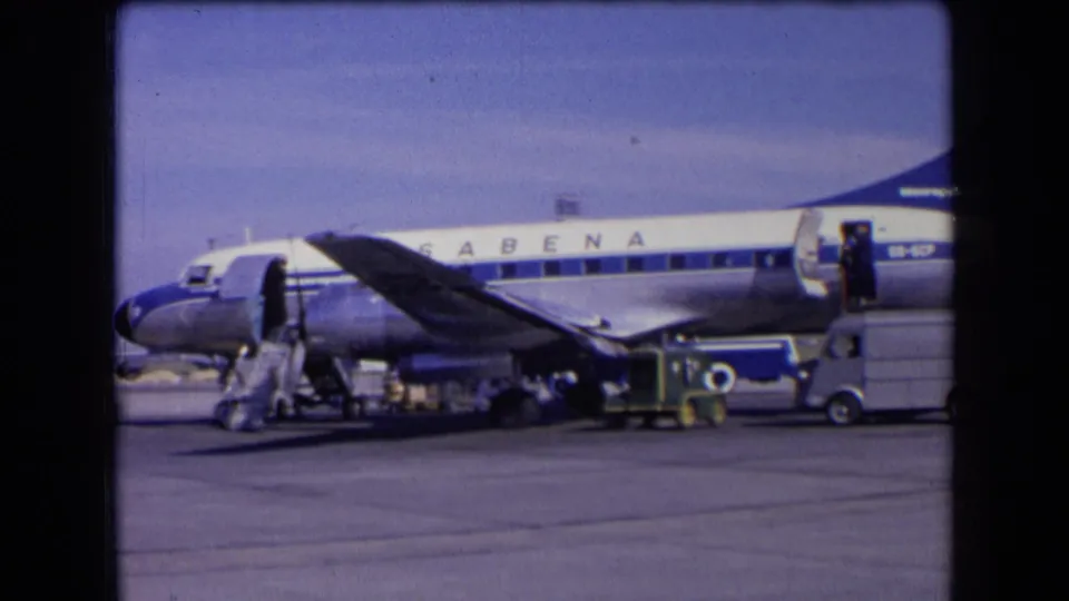 1963 France home-movie frame showing a Sabena aircraft on the tarmac with passenger stairs, ground activity, and service vehicles.
