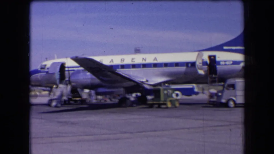 1963 France home-movie still of a Sabena aircraft on the airport apron with service equipment, people, and passenger stairs visible.
