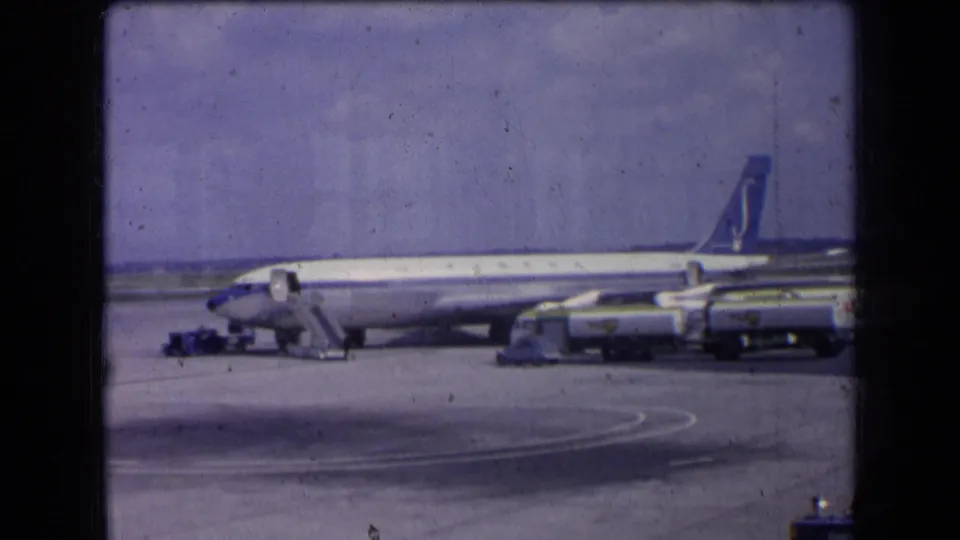 Wide 1963 France 8mm view of a passenger jet on the tarmac with mobile stairs, service vehicles, apron markings, and hazy sky.