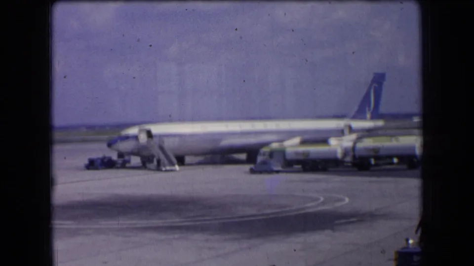 Final 1963 France frame showing a passenger aircraft parked on the apron with stairs, service vehicles, runway markings, and soft 8mm film grain.